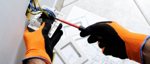Electrician wearing orange safety gloves using a screwdriver to install or repair electrical wiring in a wall outlet, illustrating essential electrical safety tips for safely handling home electrical projects.