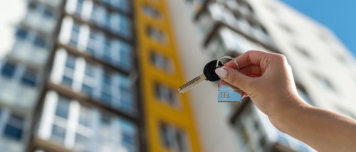 A renter's hand holding an apartment key with a small house keychain, with a modern apartment building in the background.