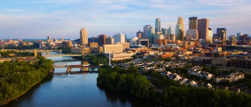 Aerial view of downtown Minneapolis, MN, with a mix of modern high-rise buildings, bridges over the river, and residential areas surrounded by lush greenery under a cloudy sky.