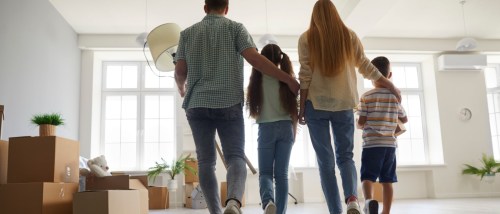 A family, including a mom, dad, and children, entering a spacious, modern, and light living room full of cardboard boxes. The image shows a backside view from behind. The scene represents a renter with children moving into their new home.