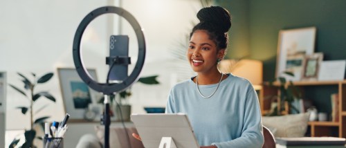 A person sits at a desk in an influencer apartment with a phone mounted on a stand equipped with a ring light and a tablet device on the desk.