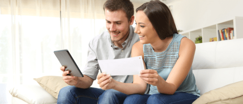 A smiling couple sits on a couch, using a tablet and holding a document to compare renters insurance policies.