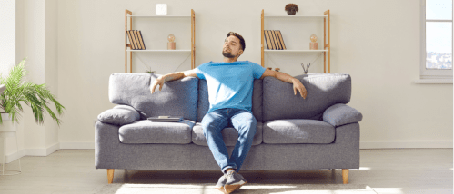 Relaxed man sitting on a grey sofa in his living room, enjoying fresh air after opening the windows, as a tip for spring home maintenance.